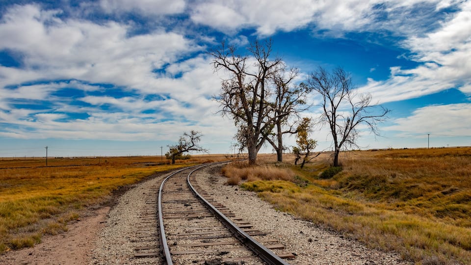 A railway track curving through a grassy field with trees and a clear sky.