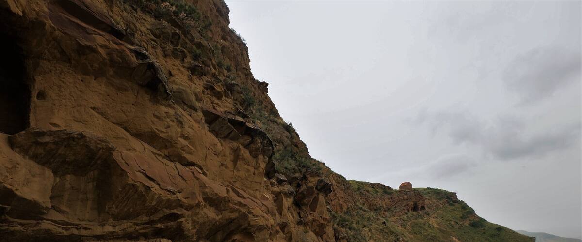 A rocky cliff with a cave entrance, a person standing on the slope, and a distant hill with a dome-shaped structure.