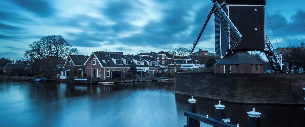 Un moulin à vent traditionnel, situé au bord d’une rivière paisible, entouré de maisons et surplombé par un ciel nuageux.