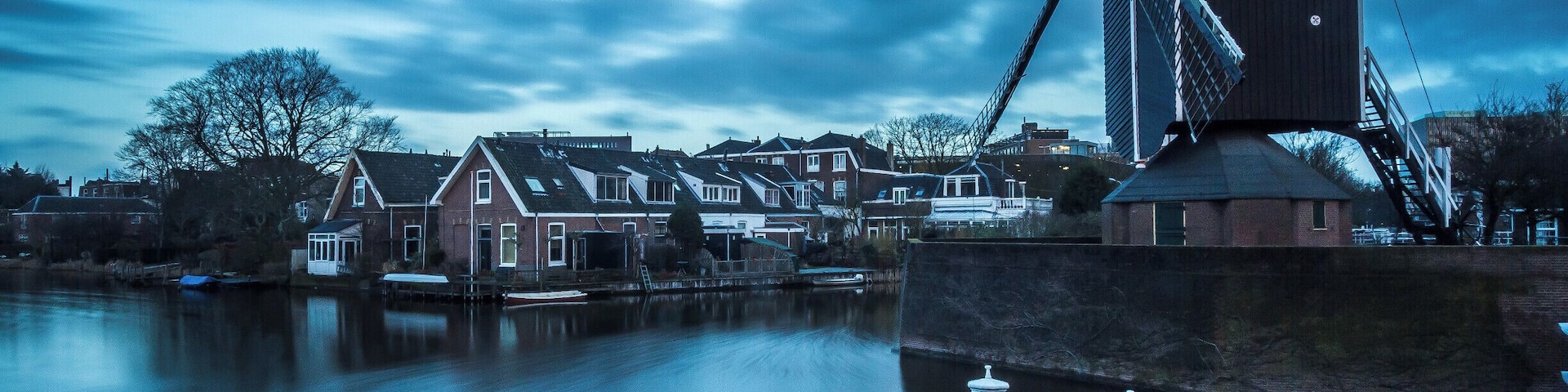 A traditional windmill by a calm river, surrounded by houses and a cloudy sky.