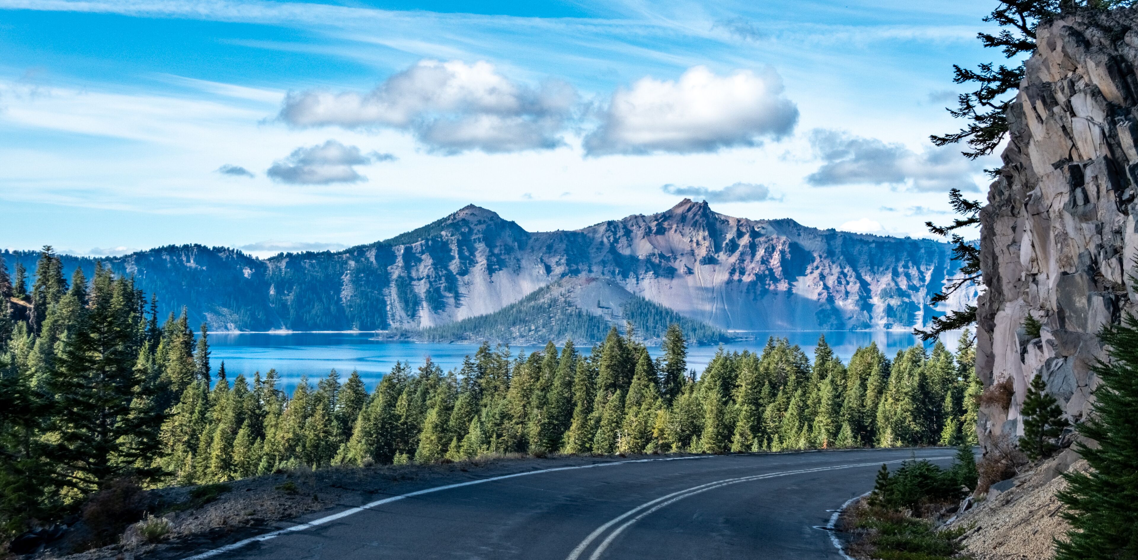 A winding road, dense coniferous forest, a mountain range, and a lake.