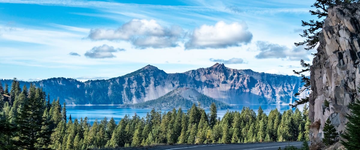 A winding road, dense coniferous forest, a mountain range, and a lake.