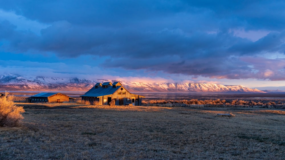 A rustic wooden barn with a blue roof, surrounded by a dry grass field and a mountainous backdrop under a cloudy sky.