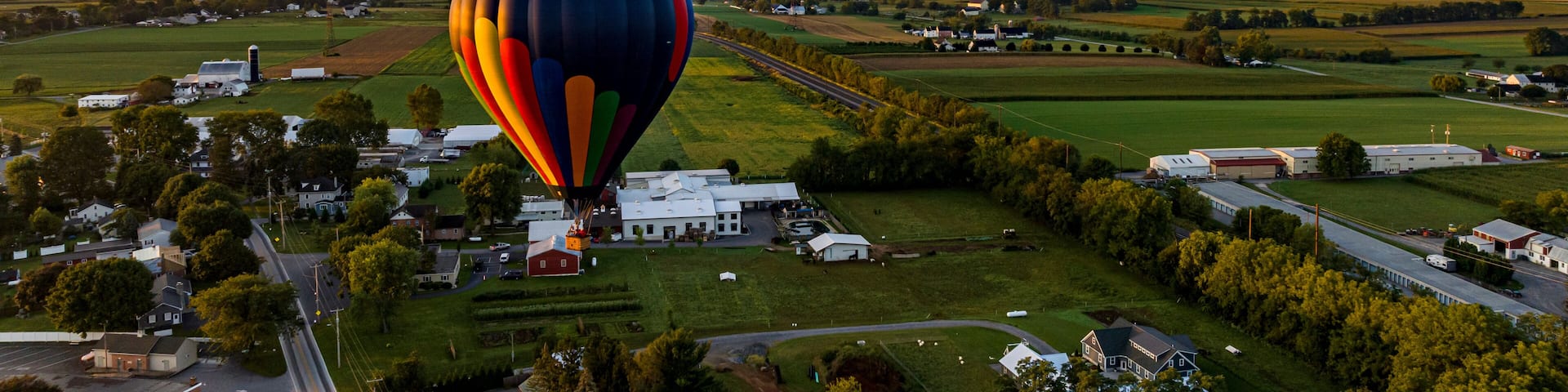 A hot air balloon over a rural landscape with fields, houses, and a road.