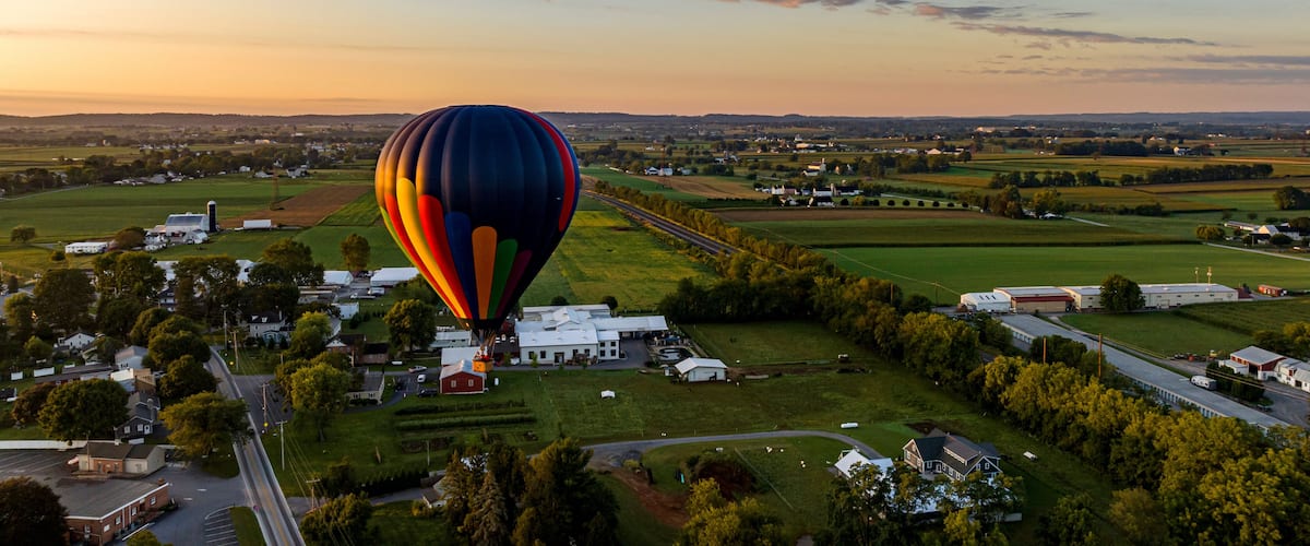 A hot air balloon over a rural landscape with fields, houses, and a road.