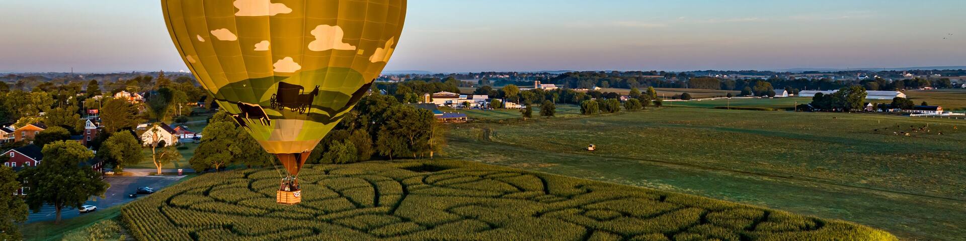 An Aerial View of a Golden Hot Air Balloon, Just Launched and Floating Across a Field With a Corn Maze, on a Sunny Summer Morning