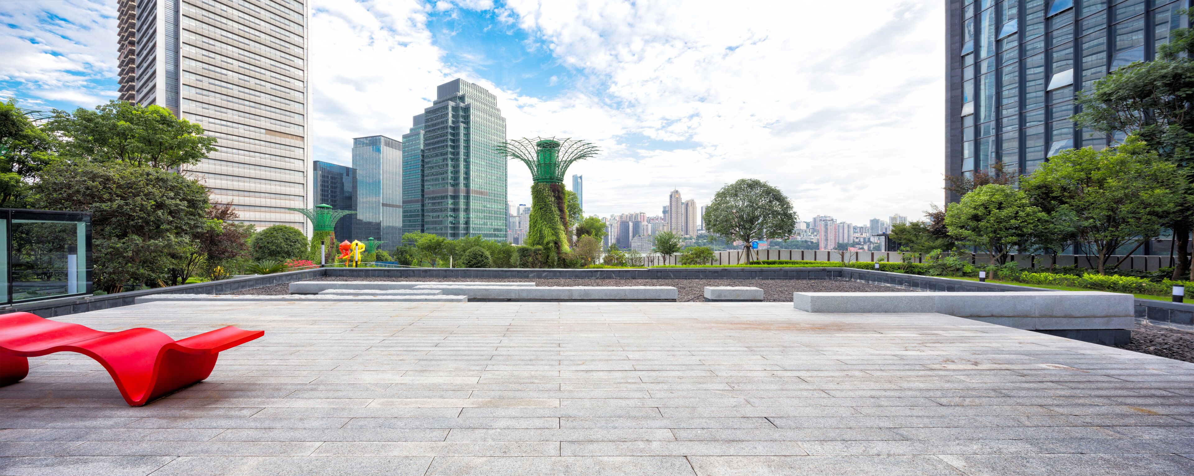 Uma praça urbana moderna com um banco vermelho, esculturas verdes e edifícios altos ao fundo.