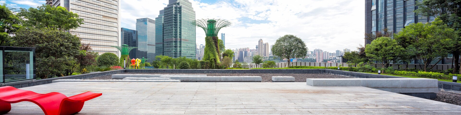 Uma praça urbana moderna com um banco vermelho, esculturas verdes e edifícios altos ao fundo.