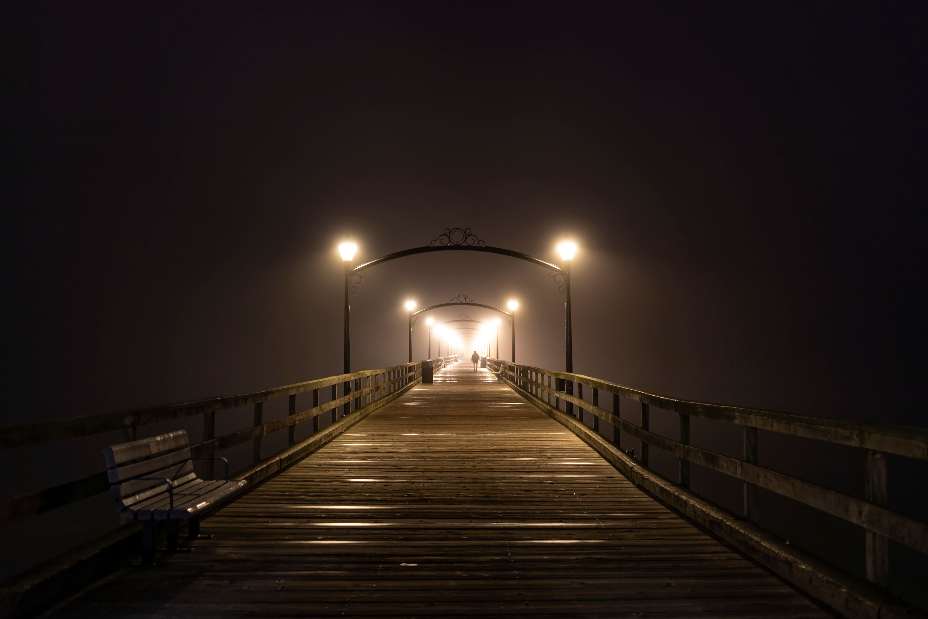 White Rock Pier illuminated with lamp light and a person in the distance taking a nighttime stroll in White Rock; British Columbia, Canada