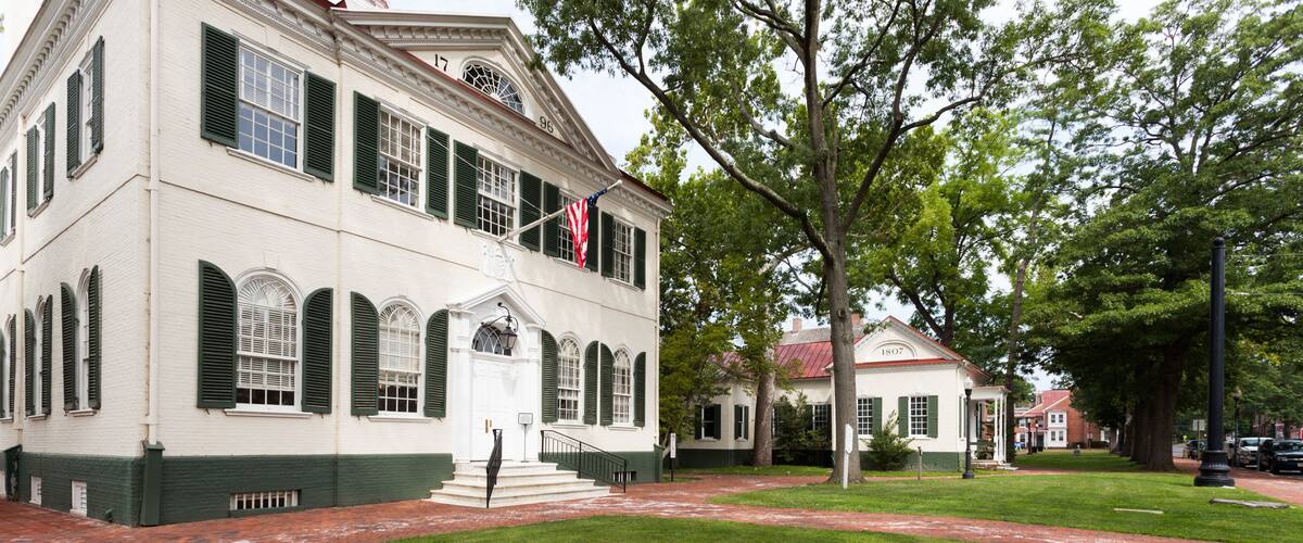 A historic white building with green shutters, a flag, and a brick pathway.