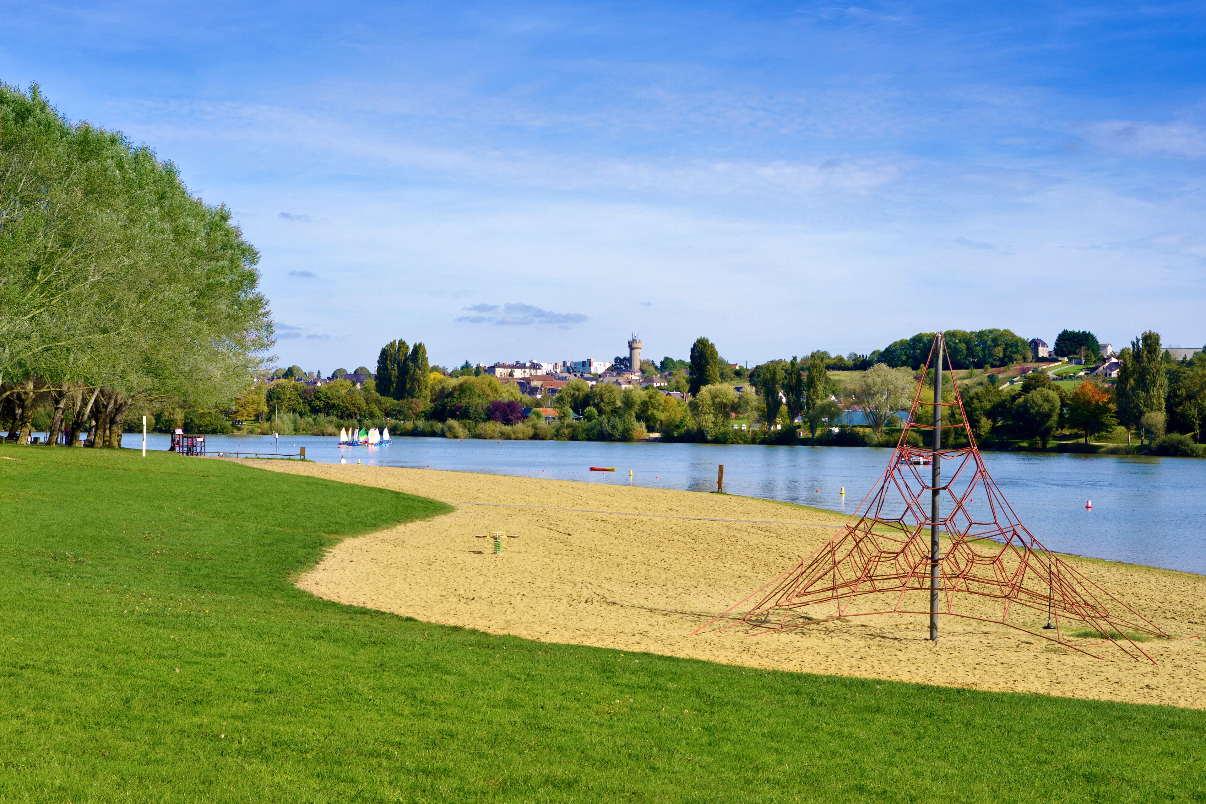 Um parque com uma área arenosa, uma estrutura de escalada vermelha e um lago com barcos.