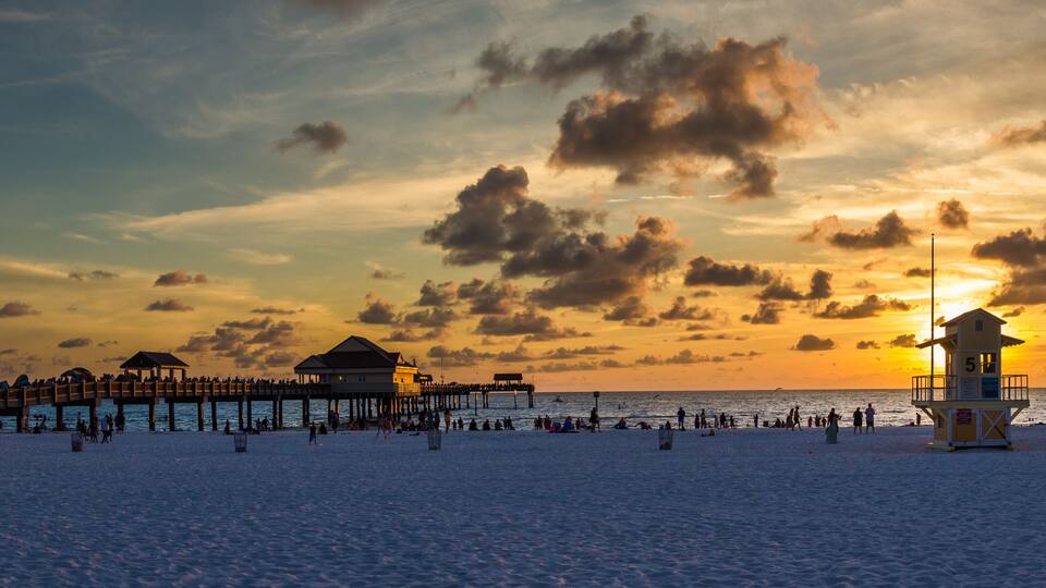 A beach at sunset with a pier, beachgoers, and a lifeguard tower.