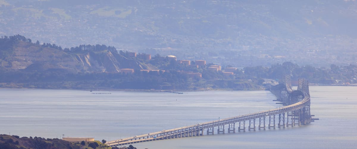 A long bridge over a calm body of water, with hills and buildings in the background.