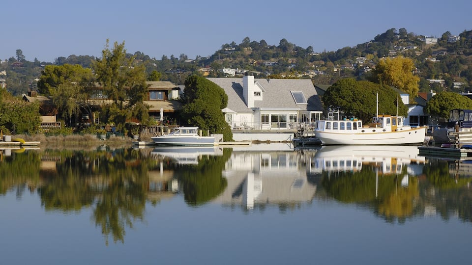 A calm lake with boats docked, a white house with a large window, and a hill with houses in the background.