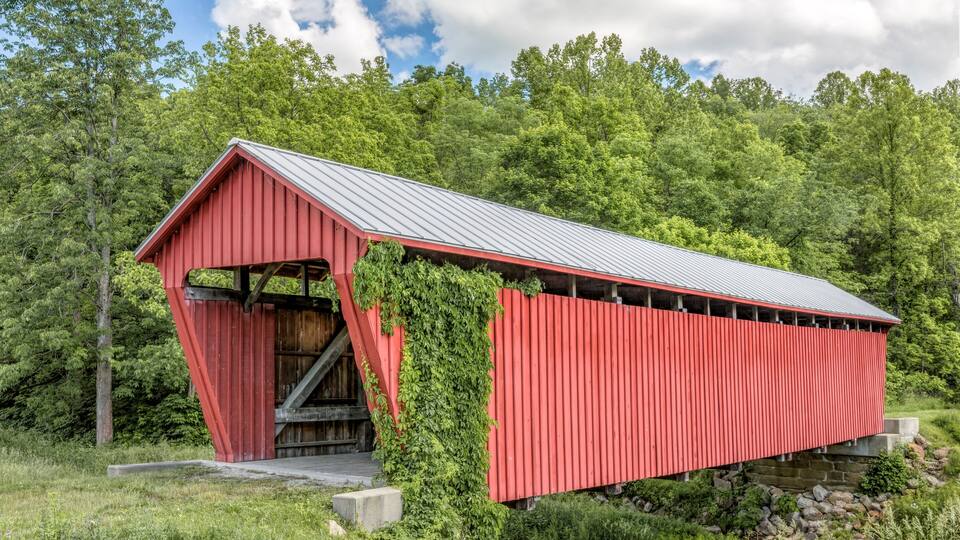 A red covered bridge with a metal roof, surrounded by lush green forest.