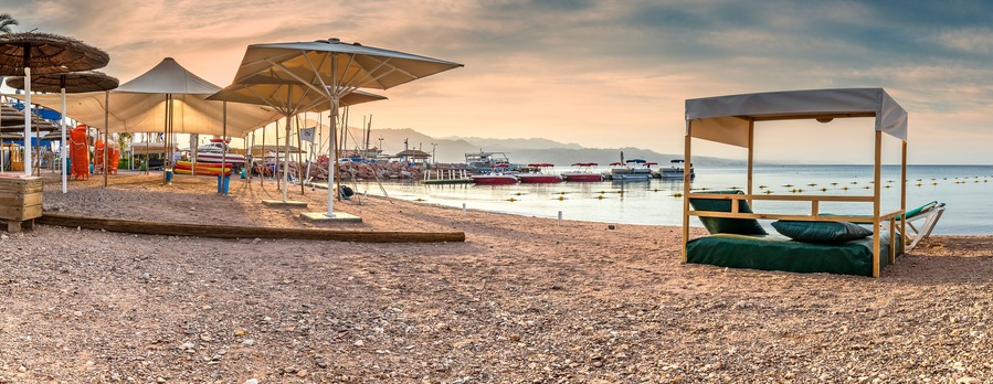 Ein Kiesstrand mit Sonnenschirmen, einer Holzliege und Booten im Hintergrund.