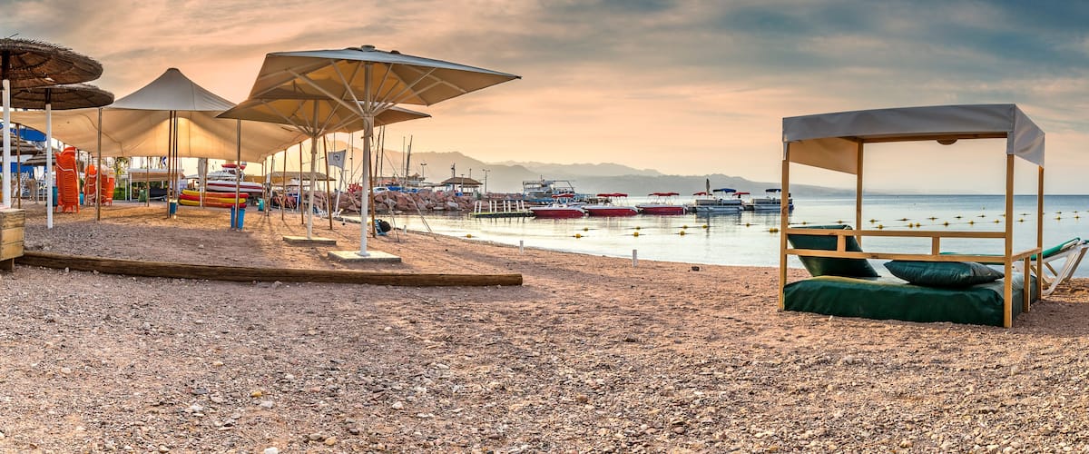 A pebbled beach with umbrellas, a wooden lounge chair, and boats in the background.
