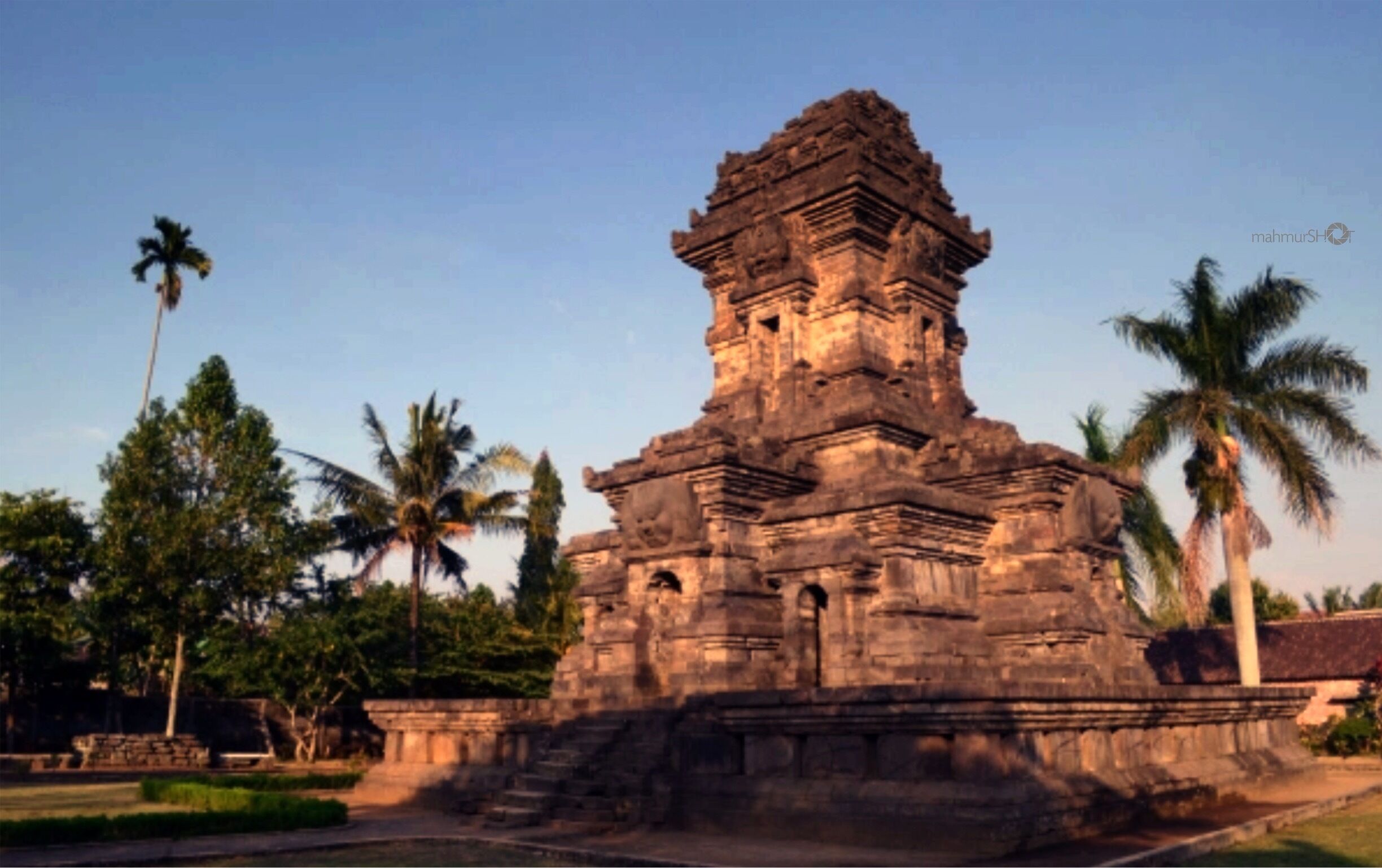 An ancient temple with intricate carvings and a tiered structure, surrounded by palm trees and greenery.