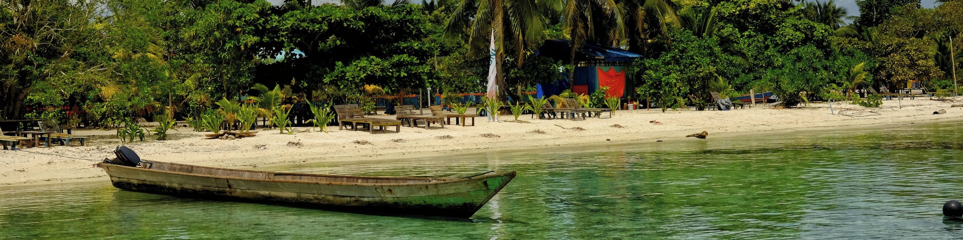 A boat on clear turquoise water, a sandy beach, palm trees, and a small structure with a red roof.
