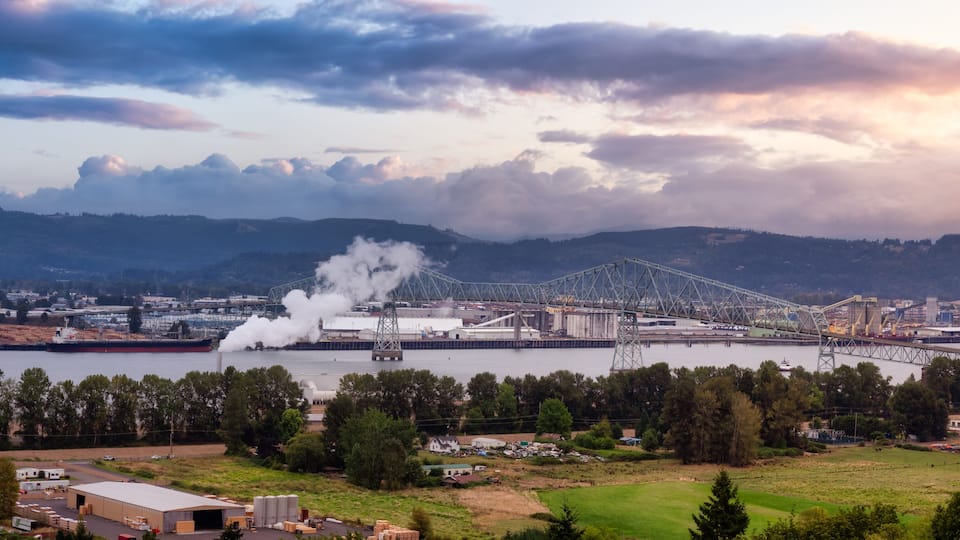 A large industrial complex with a bridge over a river, surrounded by hills and a cloudy sky.