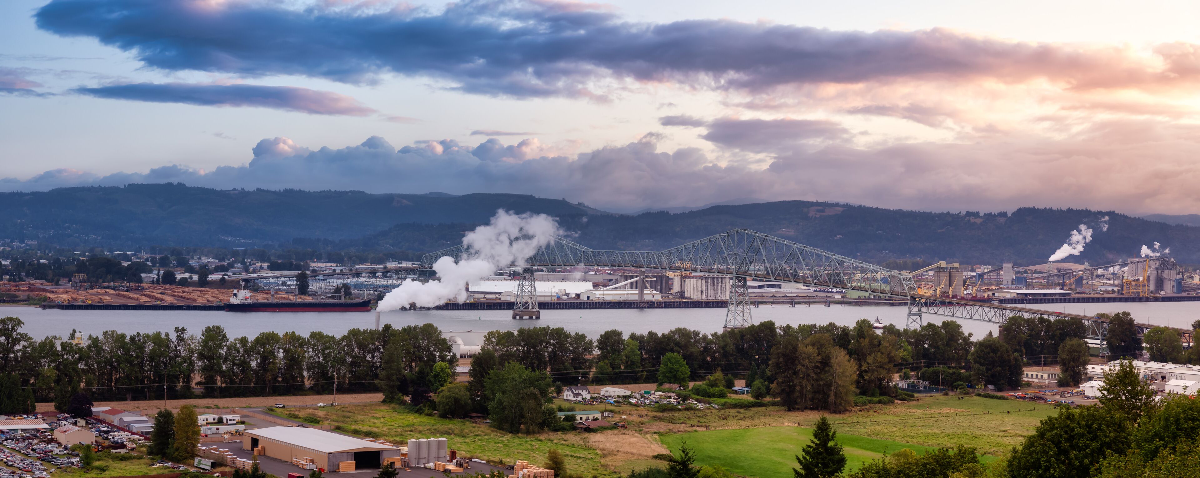 A large industrial complex with a bridge over a river, surrounded by hills and a cloudy sky.