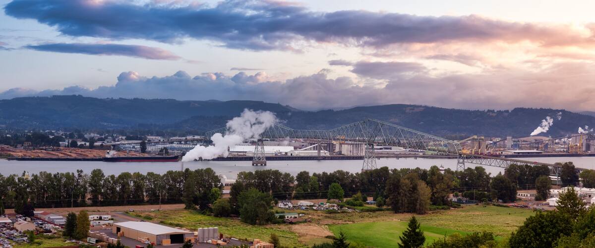 A large industrial complex with a bridge over a river, surrounded by hills and a cloudy sky.