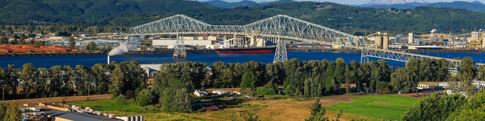 A large steel bridge spanning a river, surrounded by industrial buildings and a forested mountain range.
