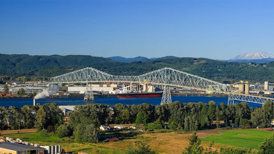 A large steel bridge spanning a river, surrounded by industrial buildings and a forested mountain range.