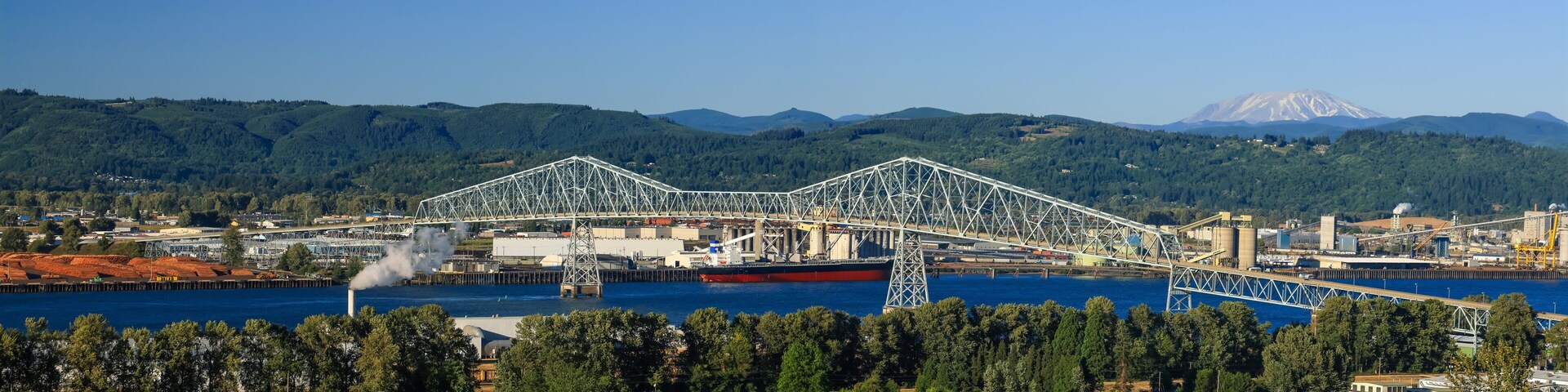 A large steel bridge spanning a river, surrounded by industrial buildings and a forested mountain range.