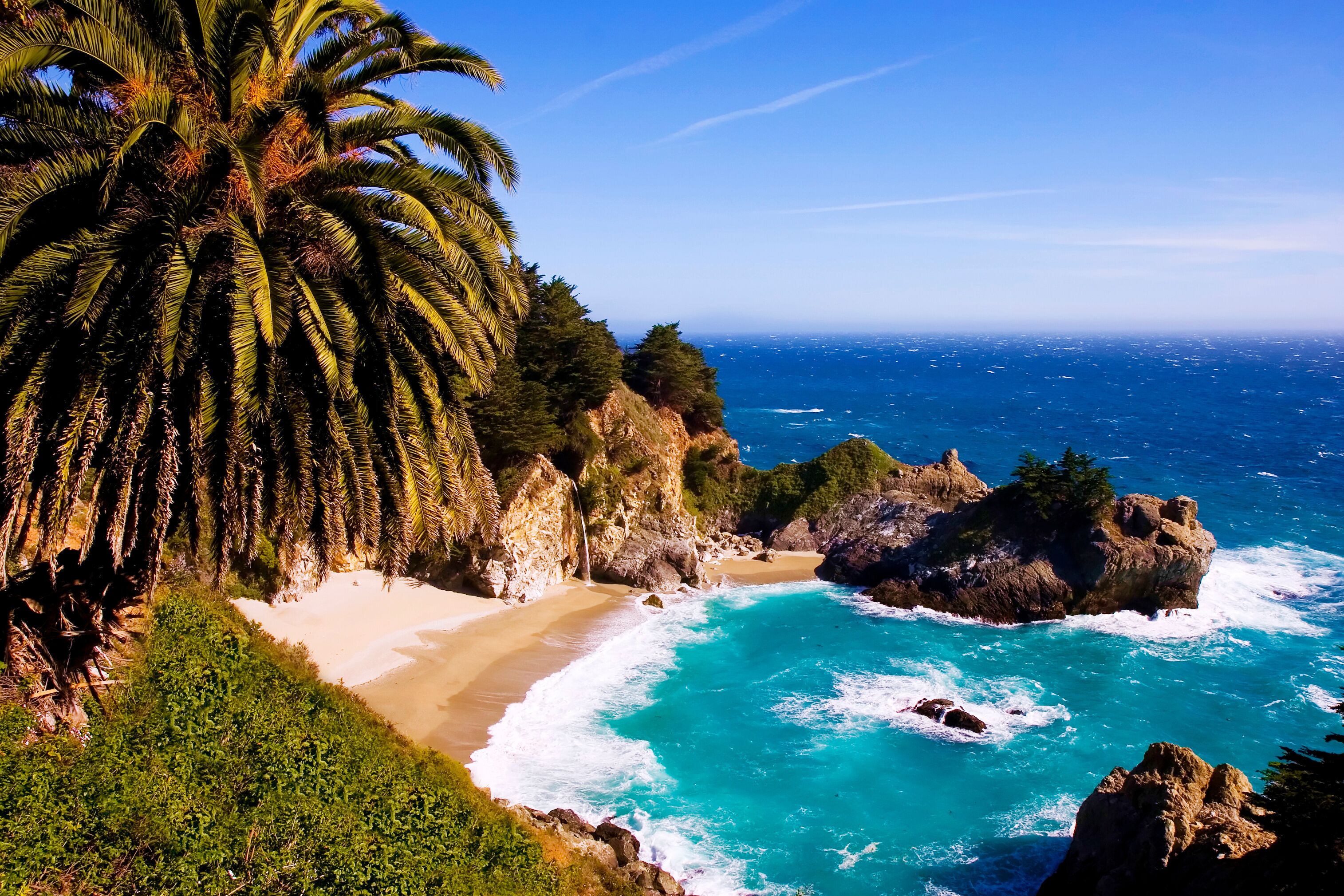 A coastal landscape with a sandy beach, turquoise waters, rocky outcrops, and a palm tree.