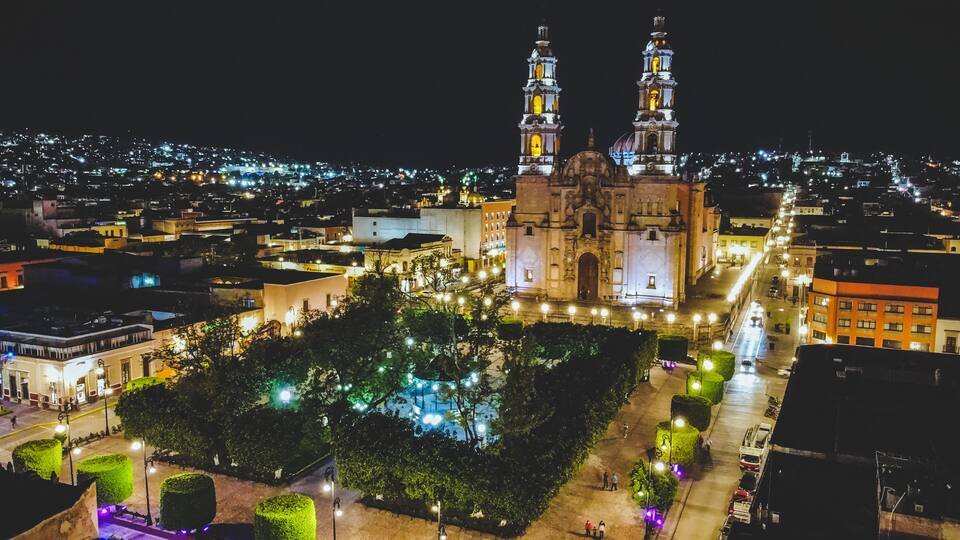 A historic cathedral with two towers, surrounded by a square with trees and street lamps.