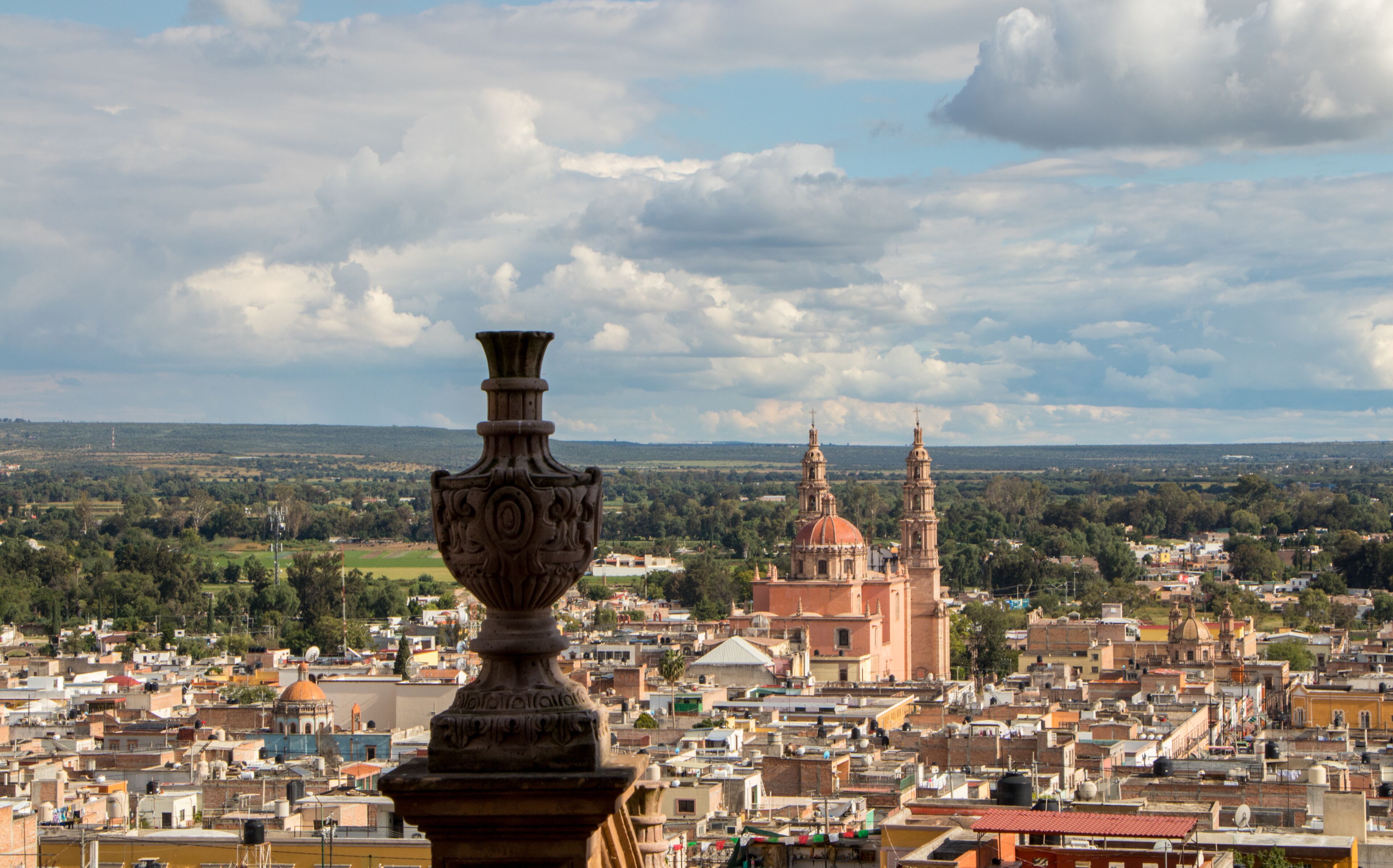 Paisagem urbana com uma torre de igreja imponente, um vaso decorativo e uma vasta área urbana sob um céu parcialmente nublado.