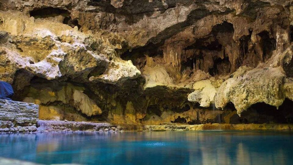 A cave with stalactites and stalagmites, a pool of water, and a cave wall.