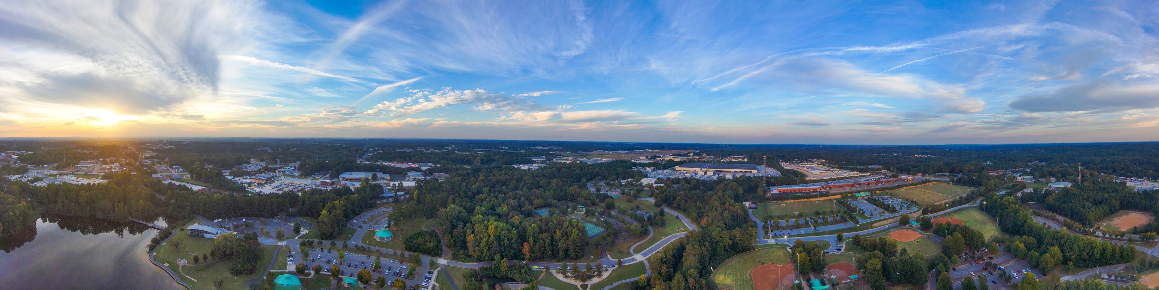 a stunning panoramic aerial shot of vast miles of lush green and autumn colored trees, still blue lake waters and blue sky with powerful cloud formations at Rhodes Jordan Park At Lawrenceville Lake in