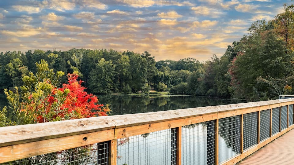 A wooden bridge over a calm lake with trees and autumn foliage in the background.