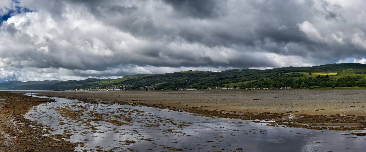 A coastal landscape with a beach, seaweed-covered shore, and distant hills under a cloudy sky.