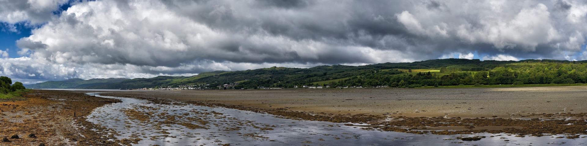 A coastal landscape with a beach, seaweed-covered shore, and distant hills under a cloudy sky.