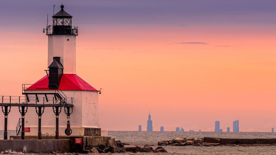 A lighthouse with a red roof near a body of water, with a city skyline in the background.