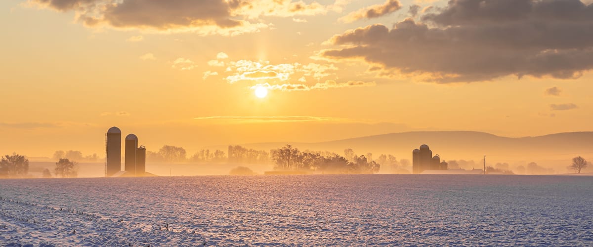 Eine verschneite Landschaft mit Sonnenaufgang, zwei Silos und einem nebelverhangenen Feld.