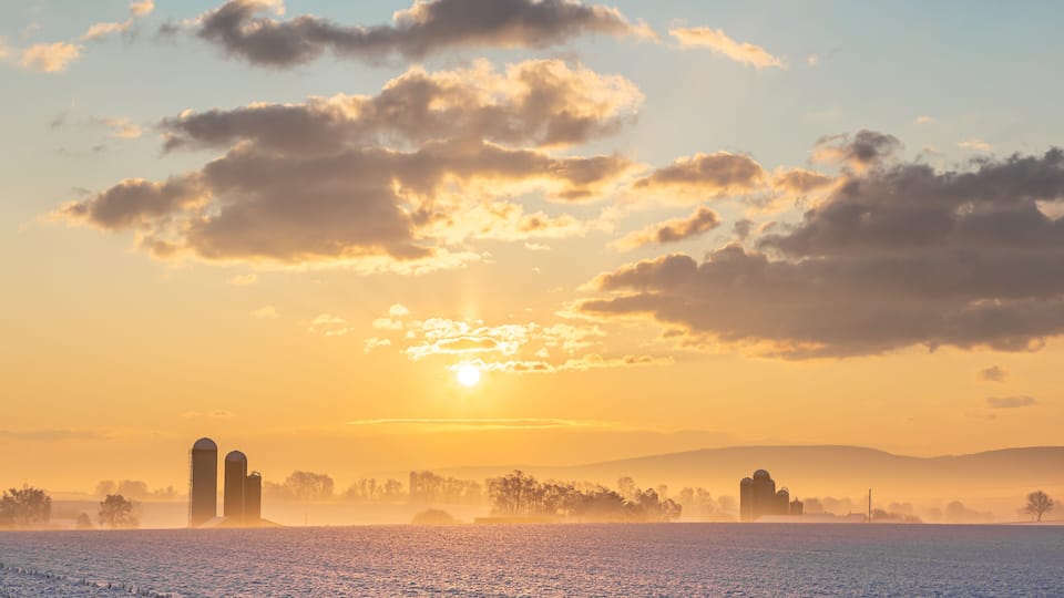 A snowy landscape with a sunrise, two silos, and a misty field.