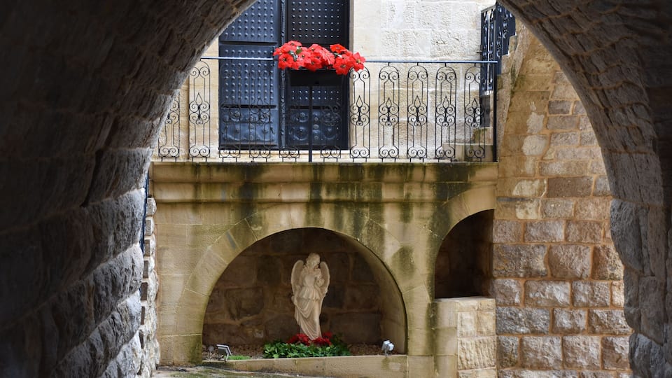 A stone archway with a statue of an angel, red flowers, and a balcony with black wrought iron railings.