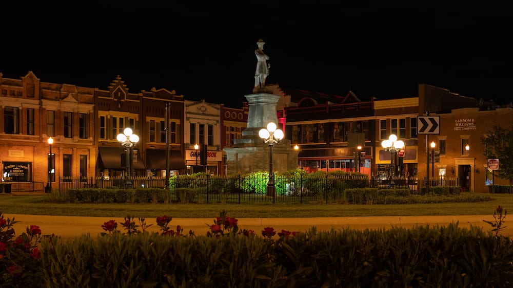 A nighttime city square with a statue, illuminated buildings, and a street sign.