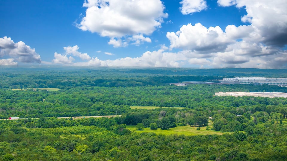 A lush green landscape with a highway, a large industrial complex, and a clear blue sky with clouds.