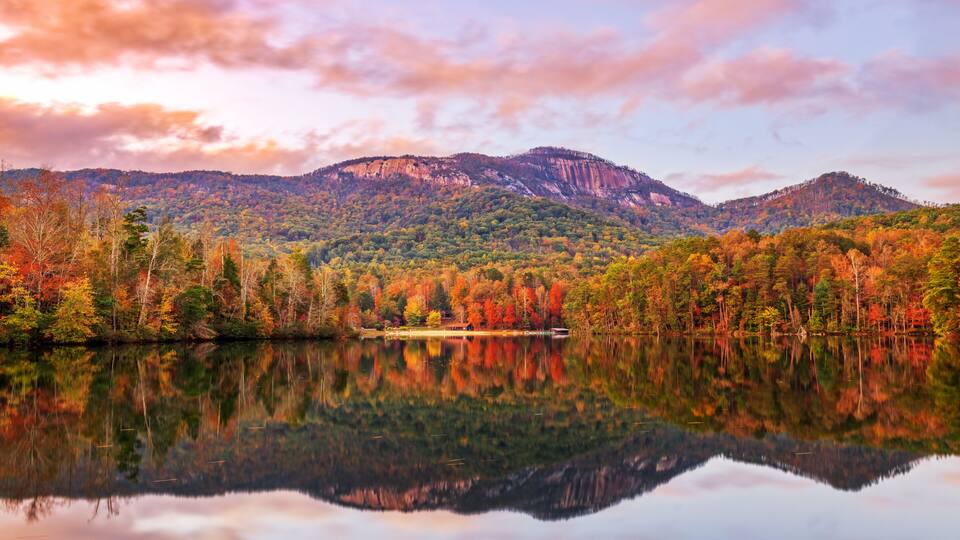 A calm lake reflecting a forested mountain range with autumn foliage.