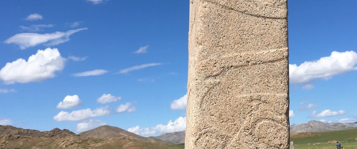 A tall, weathered stone monument with carvings, set against a clear blue sky and a mountainous landscape.