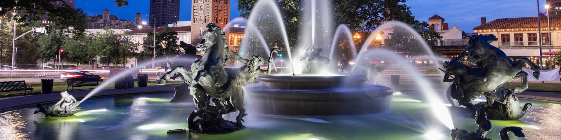 A nighttime fountain with water jets and statues, a prominent tower in the background, and surrounding trees.