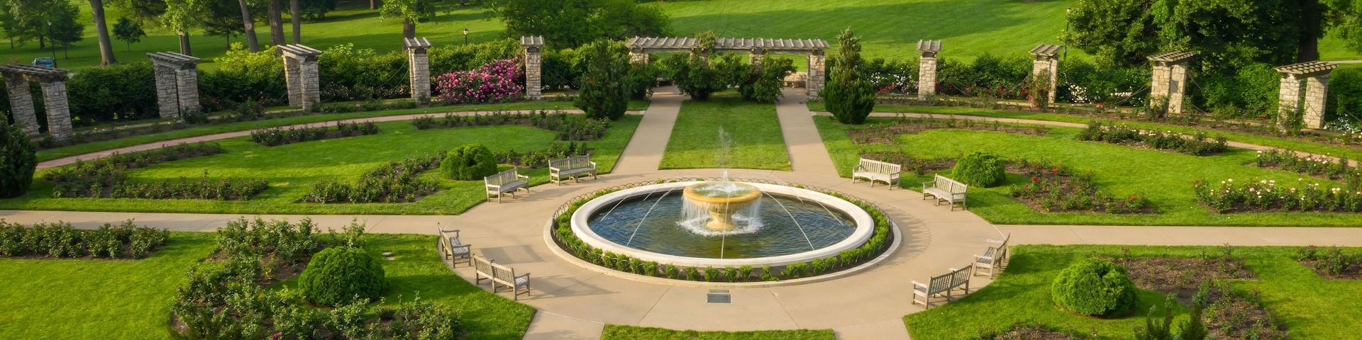 Un jardín cuidado con césped impecable, con una fuente central rodeada de pilares de piedra y bancos.
