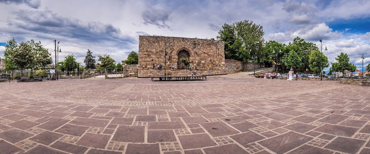 A stone building with an arched entrance, surrounded by a paved plaza with benches and trees.