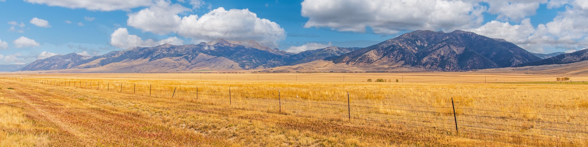 A vast open field with a fence, a mountain range in the distance, and a clear blue sky with clouds.
