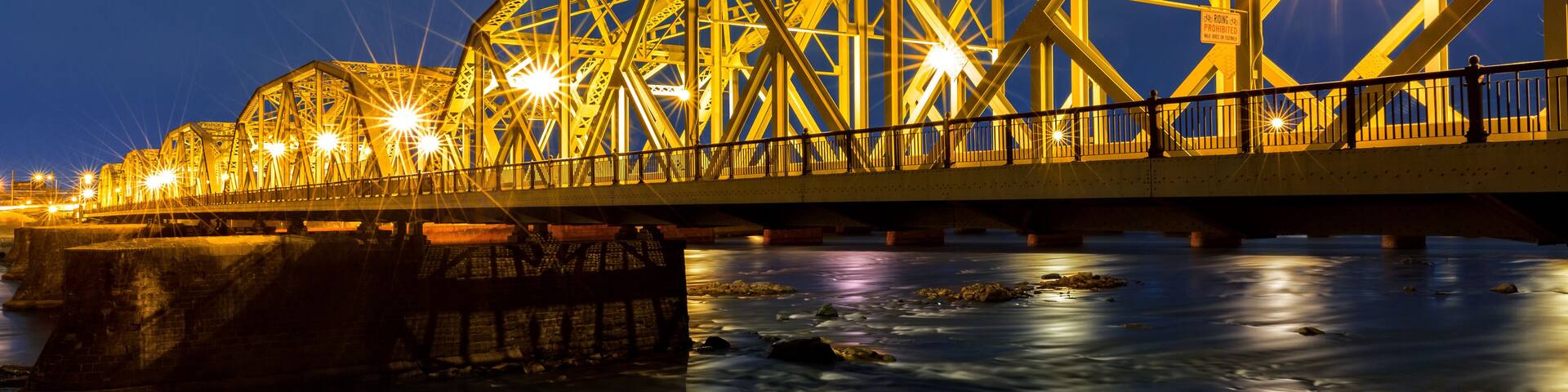 A steel bridge at night with lights reflecting on the water below.